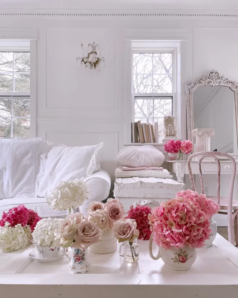 A bright room with a white sofa, various vases of pink and white flowers on a table, a vintage mirror, and a stack of books near two windows perfectly embodies Coquette Decor: How to Create a Romantic and Elegant Home.