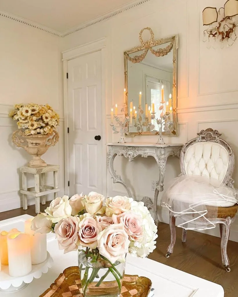 A room with an elegant vintage decor featuring a tufted chair, a table with lit candles, mirrors, floral arrangements, and a side table with flowers. Learn from Coquette Decor: How to Create a Romantic and Elegant Home to add warm lighting for a cozy ambiance.