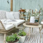 Farmhouse Summer patio with a wicker sofa and cushions, small table adorned with a vase of summer flowers, and potted plants. A decorative fence and lush greenery create the perfect backdrop for this charming outdoor seating area.