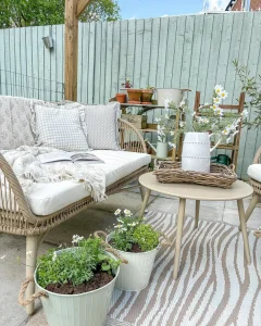 Farmhouse Summer patio with a wicker sofa and cushions, small table adorned with a vase of summer flowers, and potted plants. A decorative fence and lush greenery create the perfect backdrop for this charming outdoor seating area.