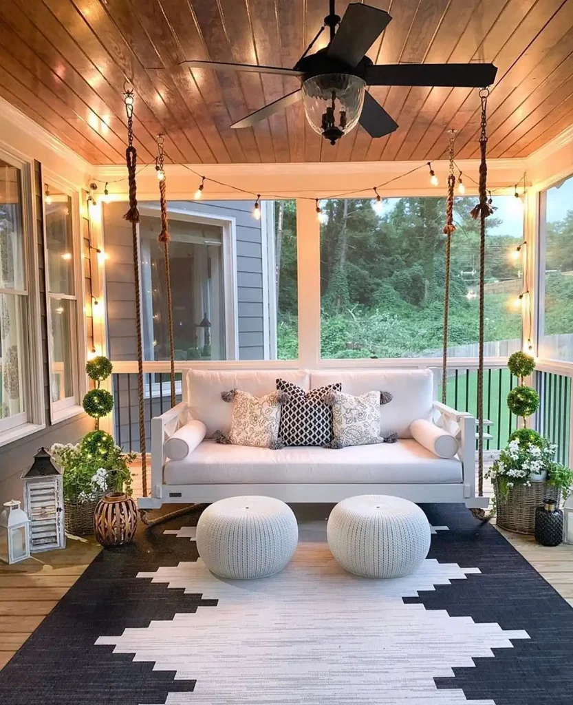 An outdoor porch with the best farmhouse porch swing for your home, two round knit poufs, string lights, potted plants, and a ceiling fan above. A black and white geometric rug adorns the wooden floor.