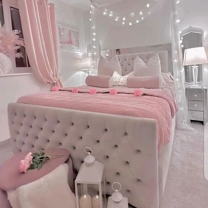 Learn How To Create A Whimsical Pink Master Bedroom Retreat with a grey tufted bed, pink bedding, and decorative pillows. The room features string lights, a mirror, bedside tables with lamps, and pink curtains. Lanterns sit beside the bed to complete the enchanting look.