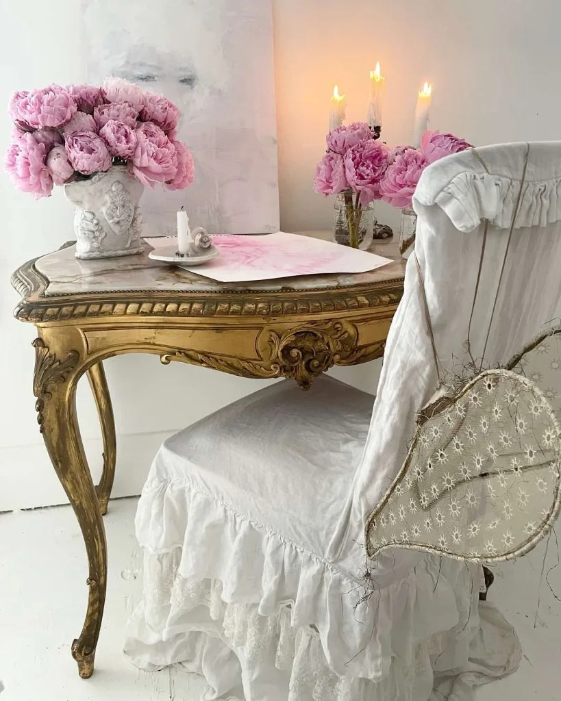 A vintage table with gold detailing is adorned with pink flowers, candles, and a painting. A white ruffled chair with lace embellishments is positioned at the table, perfect for coquette decor enthusiasts seeking to create a romantic and elegant home. Light and airy atmosphere.