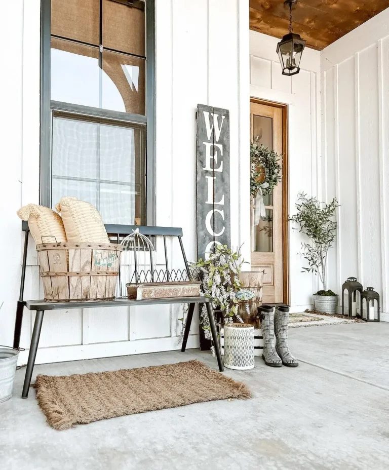 Front porch with a wooden bench, cushions, a basket, and a lantern. A "WELCOME" sign is propped up against the wall near the door. Rain boots and potted plants are placed on the concrete floor—simple tips on How to Transform Your Farmhouse Front Porch for Summer.