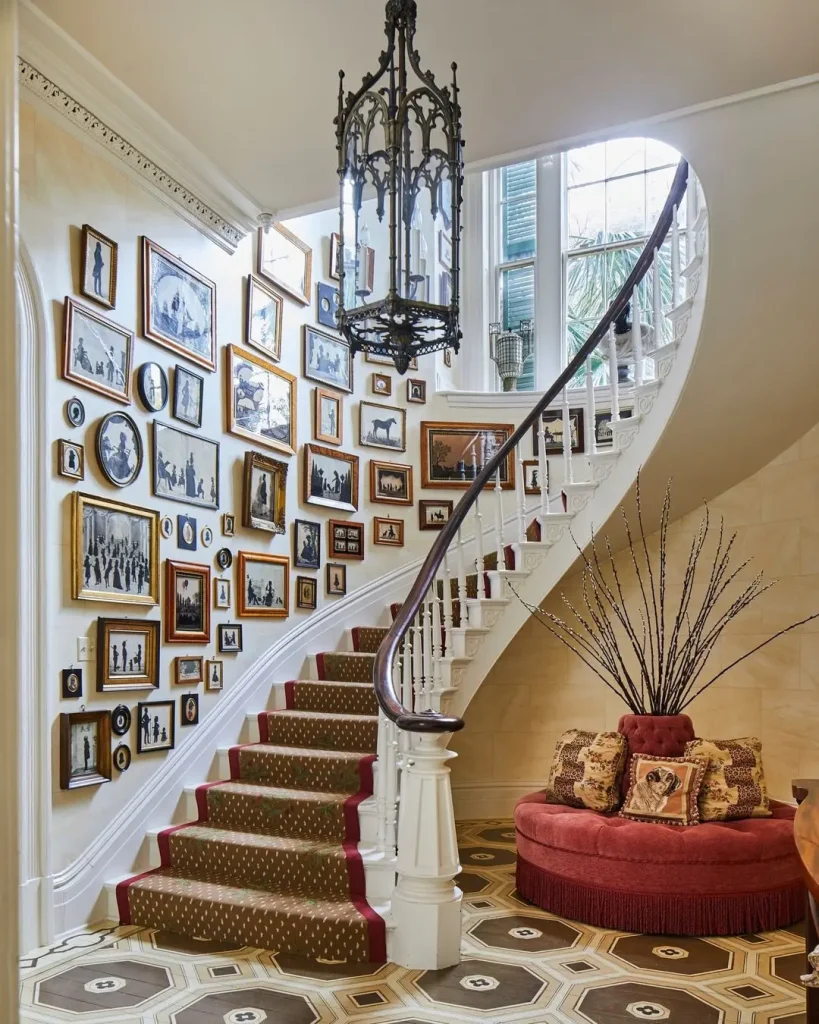 A stairway with a red carpeted path, adorned with numerous framed photos on the wall—truly a lesson in how to create a stunning gallery wall for every room. An ornate chandelier hangs above, and a round cushioned seating area sits at the base beside tall plant decor.