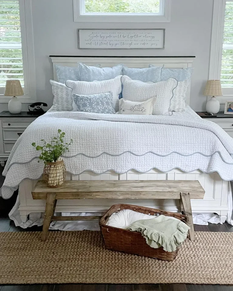 A neatly made bed with white and light blue bedding, flanked by two windows. A wooden bench sits at the foot of the bed, and a wicker basket with a blanket is on a woven rug. Two lamps are on the side tables, showcasing one of 20 Summer Bedroom Refresh Ideas That Are Affordable.