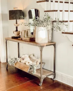 A wooden console table with metal legs showcases modern farmhouse fall decor: how to elevate your entryway with a lamp, mirror, potted greenery, and decor. The lower shelf displays a wooden crate filled with pumpkins and cozy pillows.