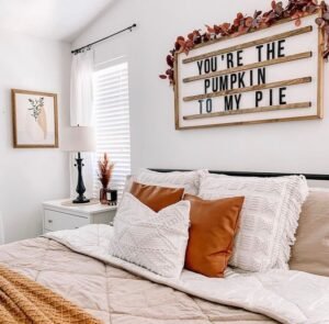 Neutral-toned bedroom with a made bed, throw pillows, a nightstand with a lamp, and a cozy wall sign reading "YOU'RE THE PUMPKIN TO MY PIE"—a perfect example of how to make your bedroom romantic this fall.