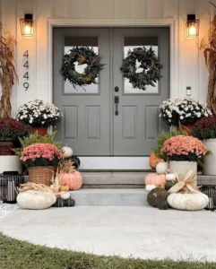 How to Transform your Farmhouse Front Porch for Fall: A front porch decorated with autumn wreaths, potted chrysanthemums, pumpkins, and corn stalks flanking a gray double door under two lantern lights.