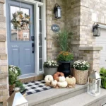 A porch with a gray door, fall wreath, lanterns, and potted plants features one of the best fall doormats to make your porch pop this season. White and orange pumpkins are arranged on the patterned mat. House number 109 is visible.