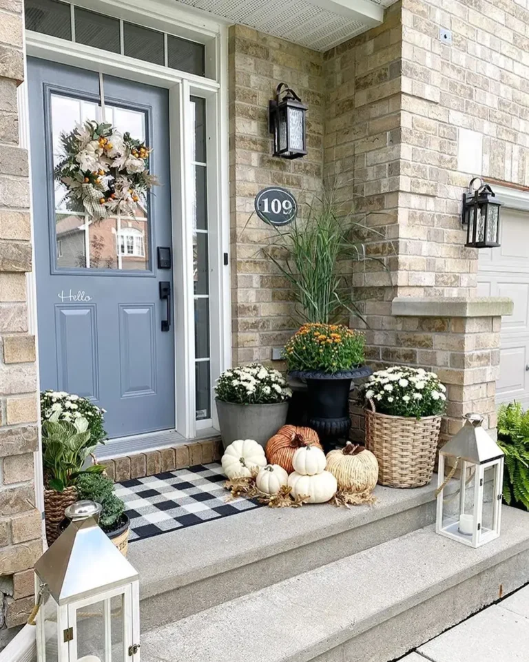 A porch with a gray door, fall wreath, lanterns, and potted plants features one of the best fall doormats to make your porch pop this season. White and orange pumpkins are arranged on the patterned mat. House number 109 is visible.