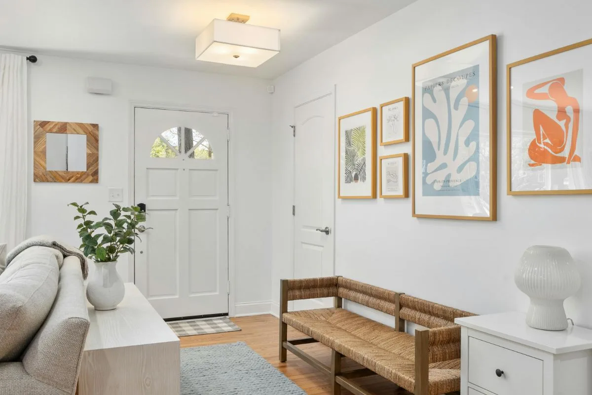 Bright entryway with a white door, natural wood bench, framed artwork on the wall, potted plant, and a white lamp on a small table—discover trendy home hacks to refresh your space with style and function.