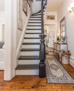 A wooden staircase with black handrail, ornate newel post, and framed art leads up from a hallway featuring a patterned rug, console table, decorative accents, and small entryway furniture chosen to fit perfectly.