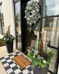 Two tall planters with winter greenery and birch branches flank a glass door adorned with a frosted wreath, atop a checkered mat and a “home is where the heart is” doormat—one of the best winter doormats to refresh your porch décor.