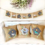 Summer farmhouse decor with burlap pillows and garland featuring buffalo check birdhouses and birds, displayed on a white bench with a floral wreath above