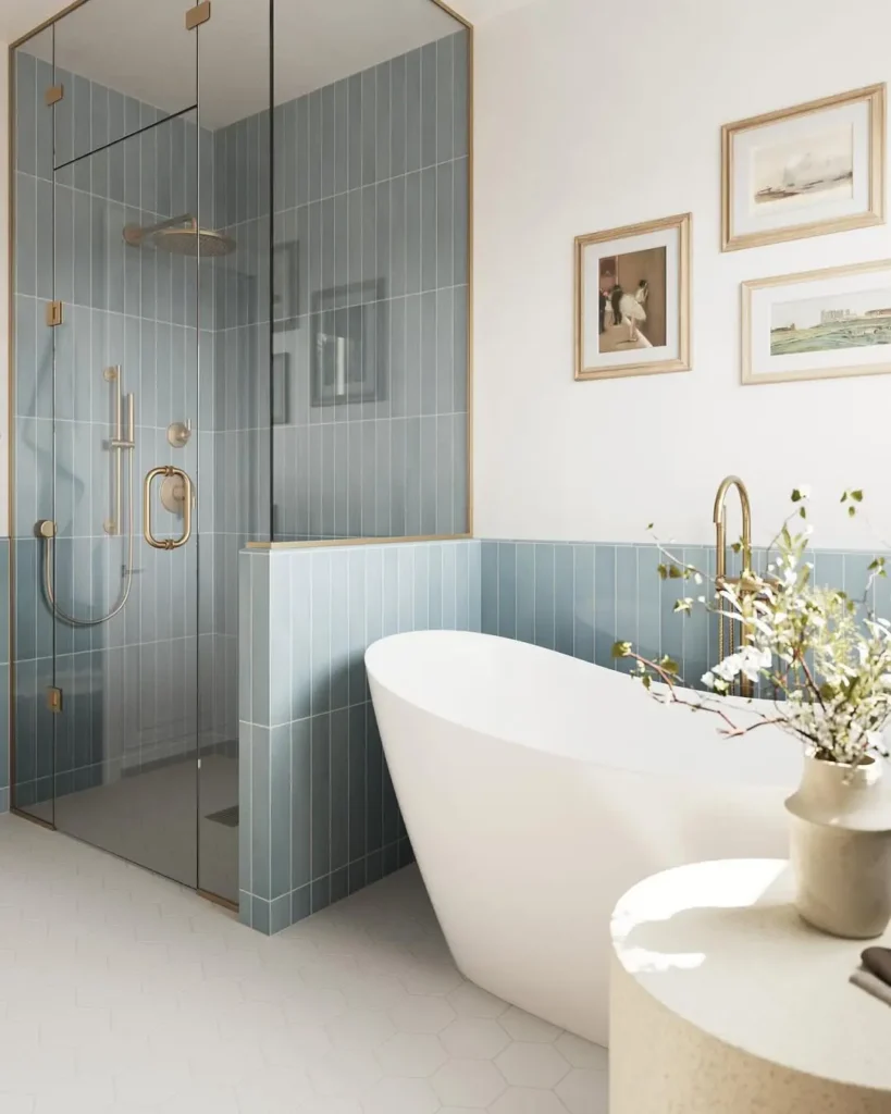 Modern coastal-inspired bathroom with a glass-enclosed shower, freestanding white bathtub, gold fixtures, blue tile accents, and framed art on the wall.