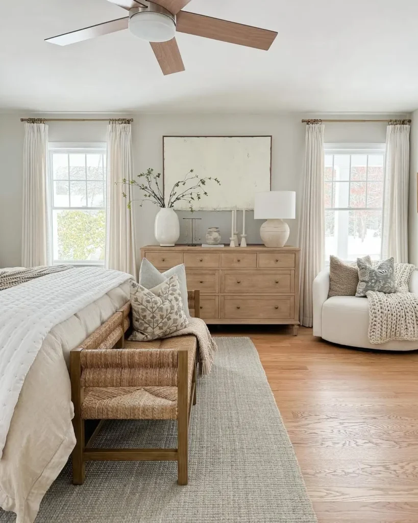 A neutral-toned bedroom with a wooden bed, dresser, armchair, and ceiling fan. Large windows let in natural light; minimal decor—including vases and a knit throw—brings a touch of serenity, perfect for adding coastal decor to any bedroom.