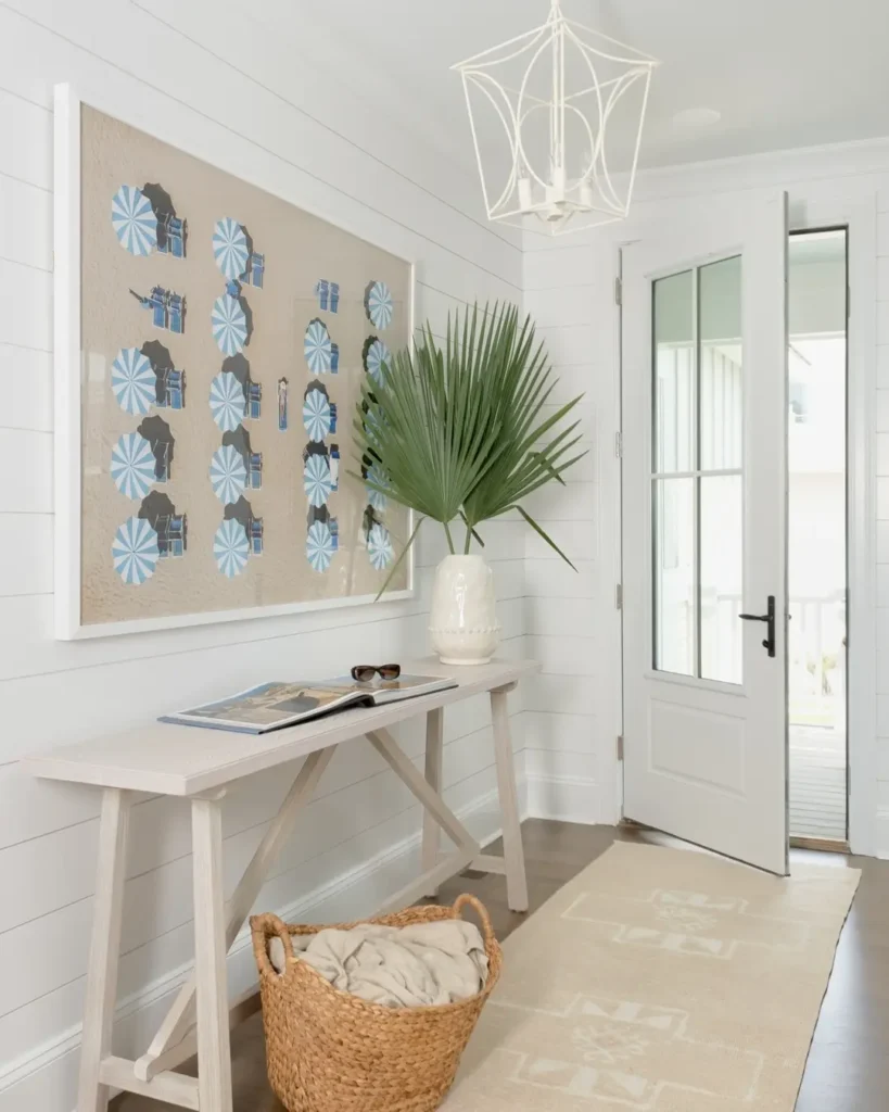 A bright entryway with a white console table, large vase holding palm leaves, wall art, woven basket, runner rug, and an open glass door letting in natural light—perfect coastal decor entryway inspiration.
