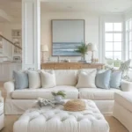 A bright living room with a white sofa, light blue and cream pillows, a tufted ottoman, and coastal decor illustrates how adding coastal decor to every room creates a serene vibe, highlighted by a large ocean painting on the wall.