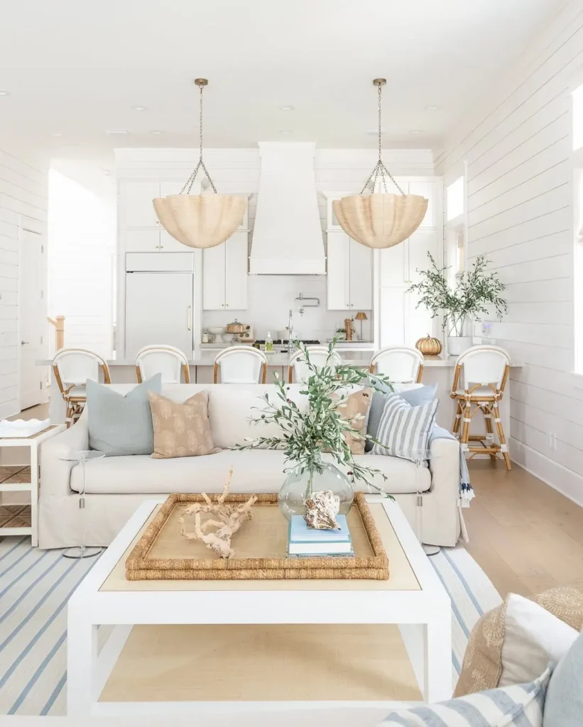 Bright, coastal-style living room and kitchen with light wood floors, white furniture, striped rug, wicker accents, pendant lights, and decorative plants make this space the perfect coastal living room to relax in.