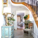 A French Country entryway features a curved staircase with ornate wrought iron railings above a hallway with a blue decorative chest, potted plants, a wicker chair, and a view into a sunlit living room.