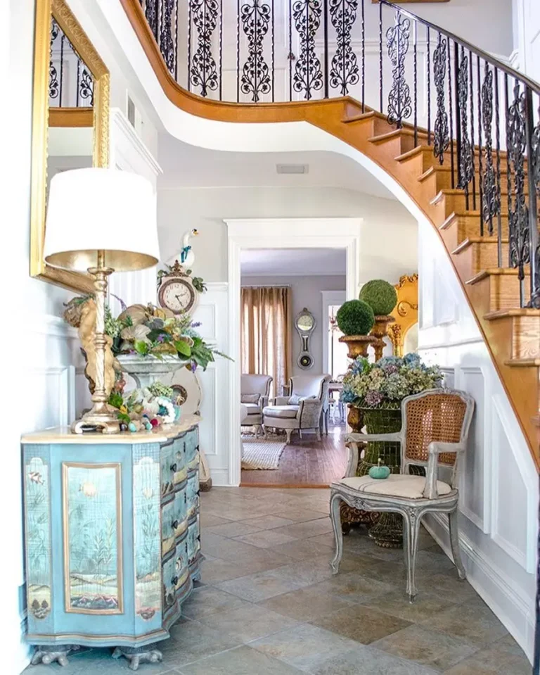 A French Country entryway features a curved staircase with ornate wrought iron railings above a hallway with a blue decorative chest, potted plants, a wicker chair, and a view into a sunlit living room.