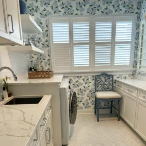 Bright laundry room with floral wallpaper, white cabinets, a marble countertop, washer, sink, open shelves, blue chair, and shuttered windows letting in natural light—showcasing one of the charming wallpaper designs for laundry room interiors.