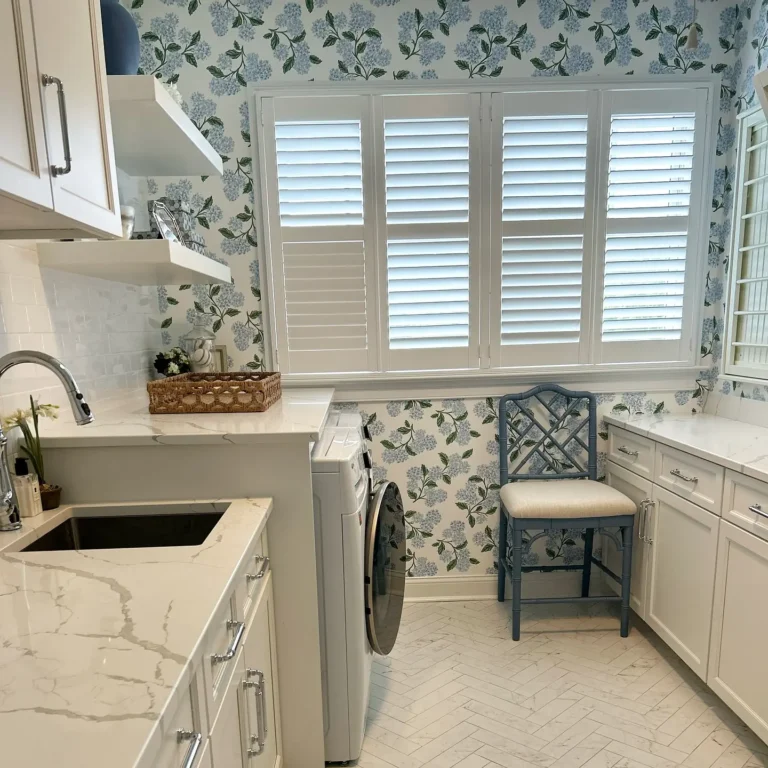 Bright laundry room with floral wallpaper, white cabinets, a marble countertop, washer, sink, open shelves, blue chair, and shuttered windows letting in natural light—showcasing one of the charming wallpaper designs for laundry room interiors.