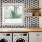 A laundry room with a large window, stylish wallpaper designs for laundry room, two front-loading machines, a sink, wooden shelves holding towels and a basket, and a wooden countertop.