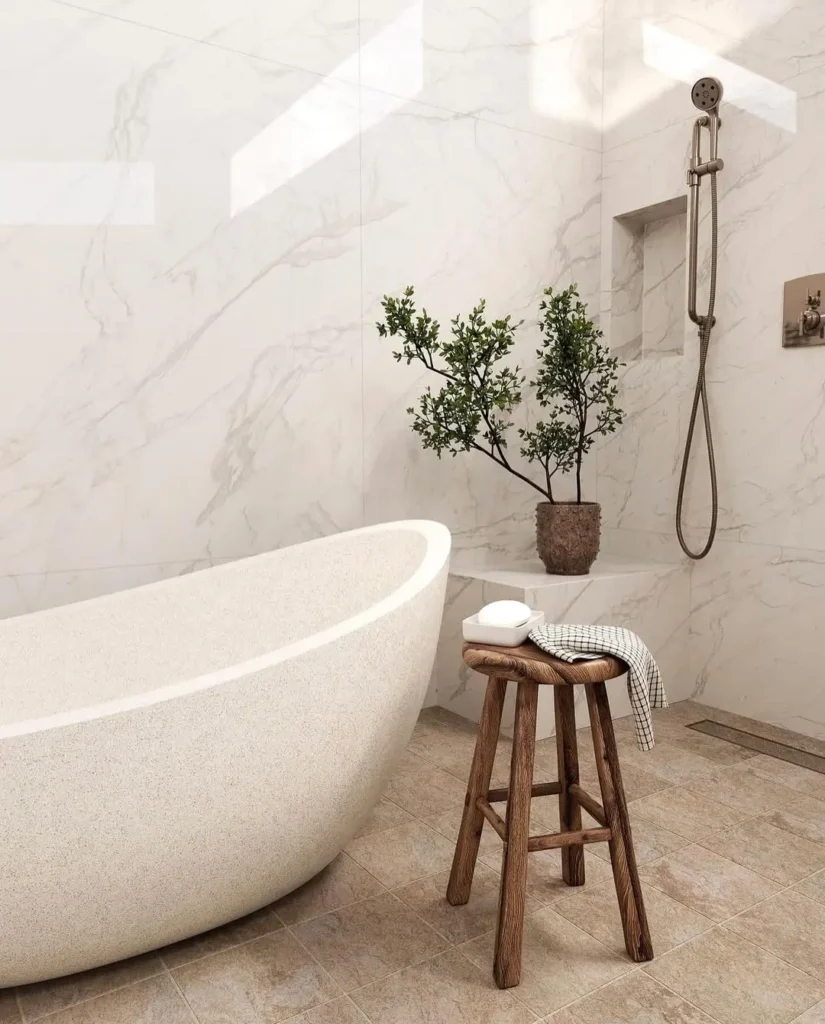 Organic Modern Bathroom with a freestanding bathtub, wooden stool holding soap and a towel, a potted plant on a bench, and a wall-mounted shower. Marble walls and beige tile floor complete the serene look.