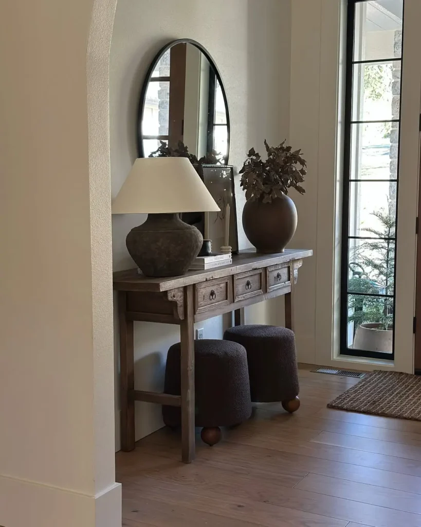 an organic modern entryway decorated with distressed looking wooden console styled with earthy vase and matching table lamp next to a wall circular mirror make this space warm and inviting