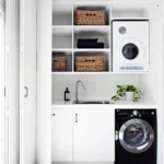 A compact laundry area features a smart laundry room layout with a white countertop, sink, potted plant, front-load washer and dryer, shelves with baskets, and neatly folded towels.