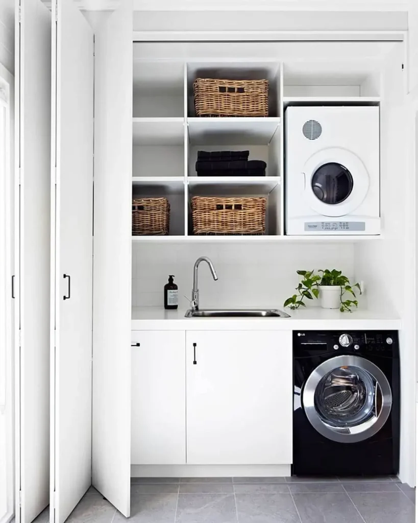 A compact laundry area features a smart laundry room layout with a white countertop, sink, potted plant, front-load washer and dryer, shelves with baskets, and neatly folded towels.