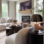 A modern living room features a light sofa, boucle chair, round coffee tables, a fireplace with art above, earthy texture in organic modern decor, and tall windows with beige curtains.