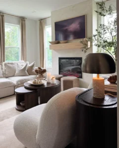 A modern living room features a light sofa, boucle chair, round coffee tables, a fireplace with art above, earthy texture in organic modern decor, and tall windows with beige curtains.