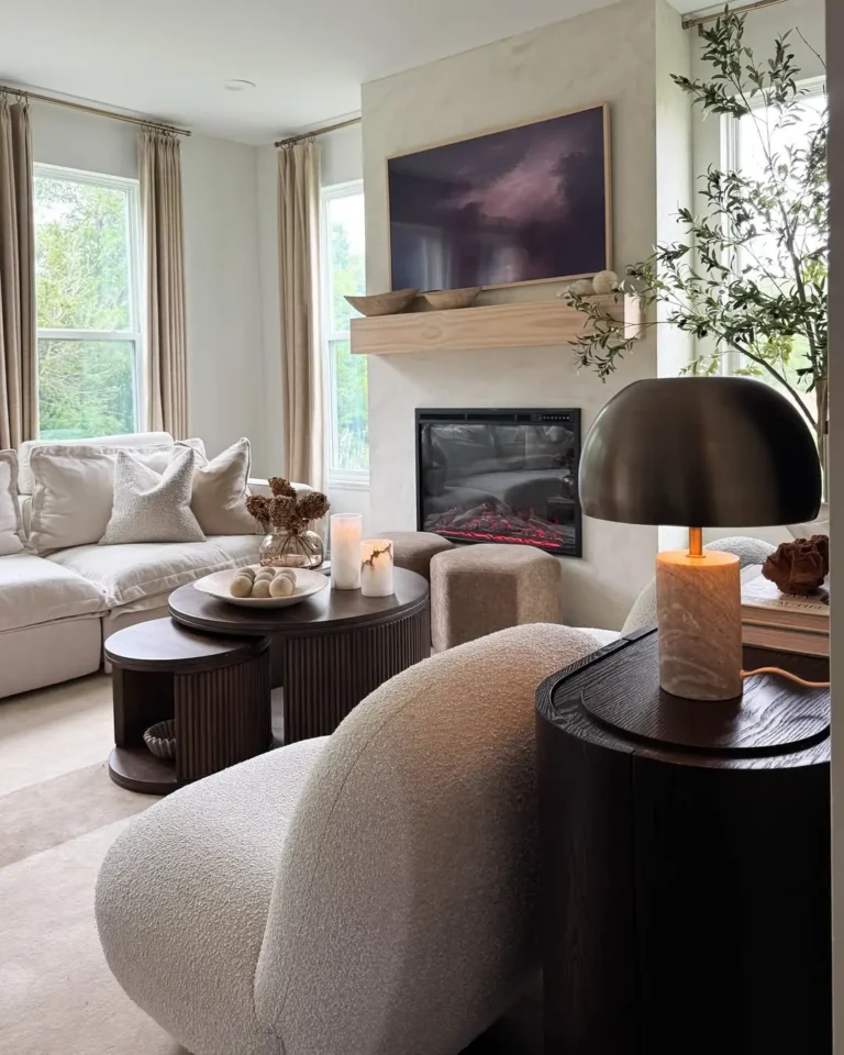 A modern living room features a light sofa, boucle chair, round coffee tables, a fireplace with art above, earthy texture in organic modern decor, and tall windows with beige curtains.