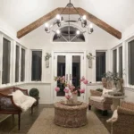 A bright sunroom with wicker furniture, a round coffee table topped with a pink floral arrangement, large windows, and a chandelier inspired by French country lighting hanging from a vaulted ceiling with exposed beams.