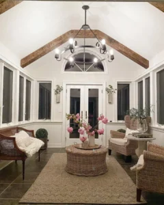 A bright sunroom with wicker furniture, a round coffee table topped with a pink floral arrangement, large windows, and a chandelier inspired by French country lighting hanging from a vaulted ceiling with exposed beams.