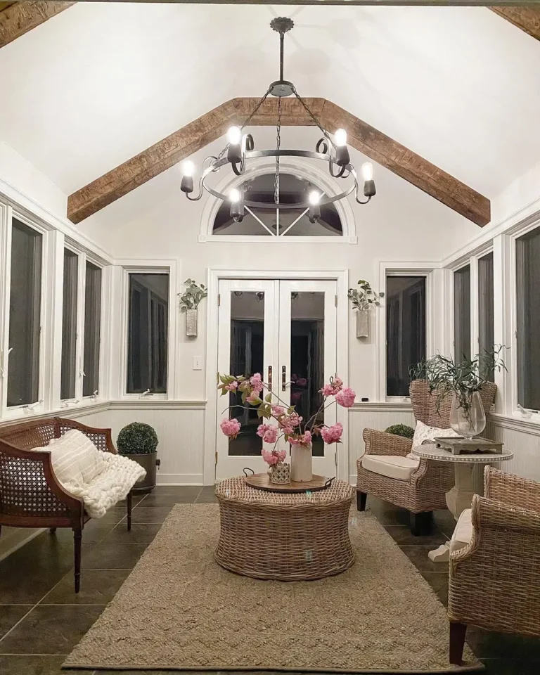 A bright sunroom with wicker furniture, a round coffee table topped with a pink floral arrangement, large windows, and a chandelier inspired by French country lighting hanging from a vaulted ceiling with exposed beams.