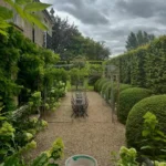 A gravel garden path lined with trimmed hedges and green plants evokes a French country backyard garden as it leads to a metal arbor and outdoor dining table under a cloudy sky.