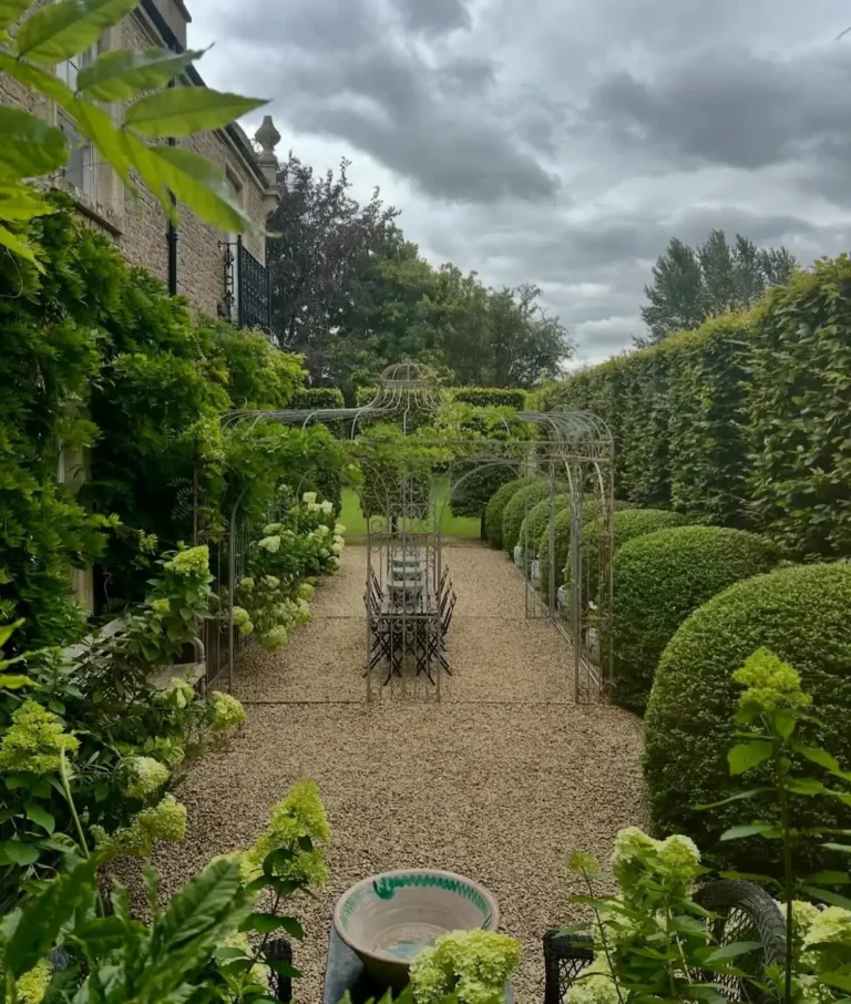 A gravel garden path lined with trimmed hedges and green plants evokes a French country backyard garden as it leads to a metal arbor and outdoor dining table under a cloudy sky.