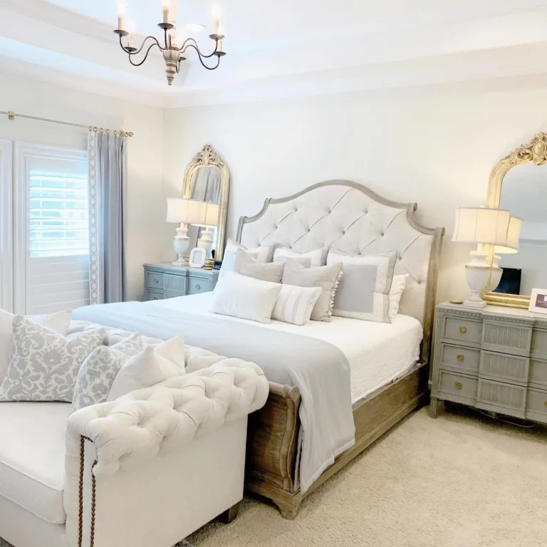 Elegant french country bedroom furniture showcasing a tufted bed, matching loveseat, two mirrored nightstands with lamps, gold-framed mirrors, and light gray curtains. The room features soft, neutral tones.