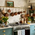 A french country kitchen with green cabinetry, a modern range beneath hanging copper pots, white tile backsplash, open shelves with plants, and a teapot on the stove.