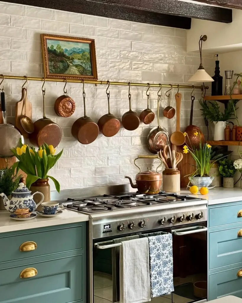 A french country kitchen with green cabinetry, a modern range beneath hanging copper pots, white tile backsplash, open shelves with plants, and a teapot on the stove.