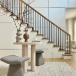 A modern interior features an Organic Modern Entryway with a small round table, two gray stools, decorative vases, and a staircase with black railings in the background.