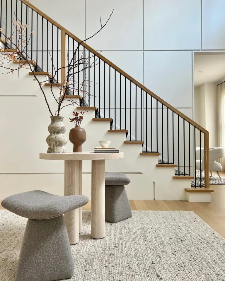 A modern interior features an Organic Modern Entryway with a small round table, two gray stools, decorative vases, and a staircase with black railings in the background.