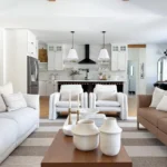Organic modern living room with neutral tones, striped rug, wooden coffee table with white vases, and an open kitchen featuring white cabinets and black accents in the background.