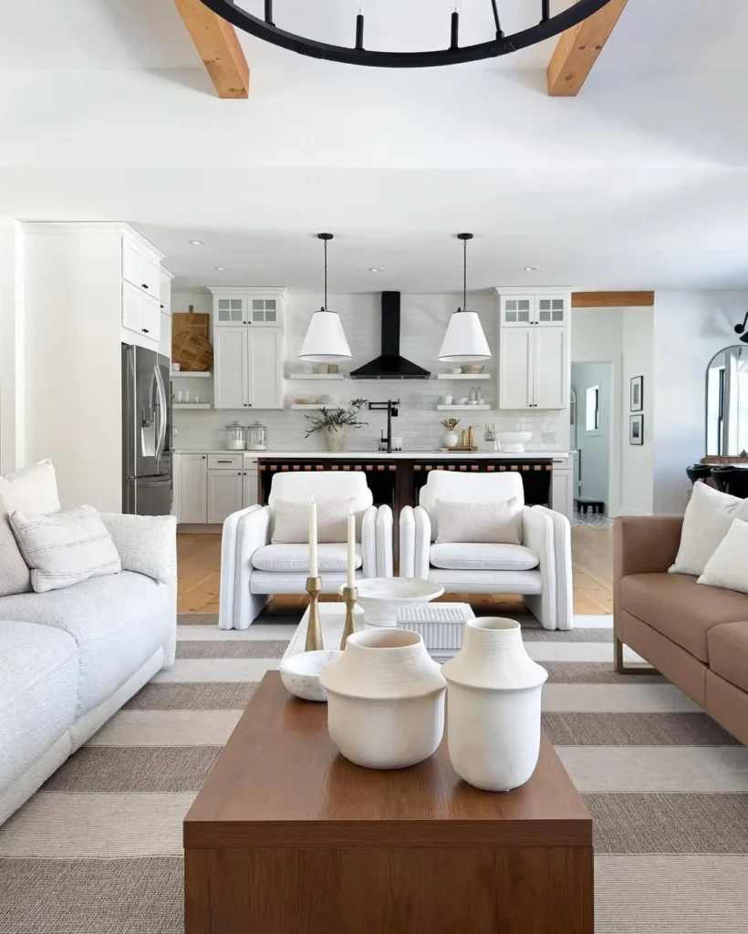 Organic modern living room with neutral tones, striped rug, wooden coffee table with white vases, and an open kitchen featuring white cabinets and black accents in the background.