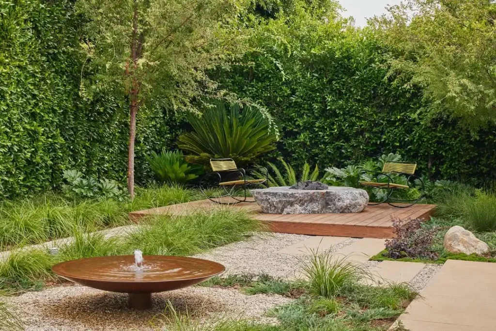 An organic modern landscaped backyard features a circular water fountain, gravel paths, a stone fire pit, wooden deck, two chairs, and lush green foliage.