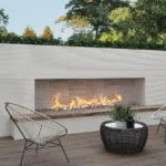 Modern organic patio with a long built-in fireplace, two wire-frame chairs, a round wicker table, and trees in the background.